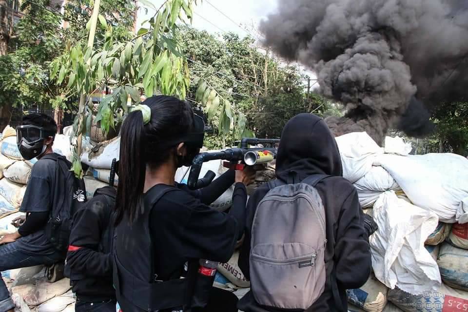 a barricade from behind in Myanmar with a youth in a gas mask looks at the camera, and a woman facing away fires a home made gun