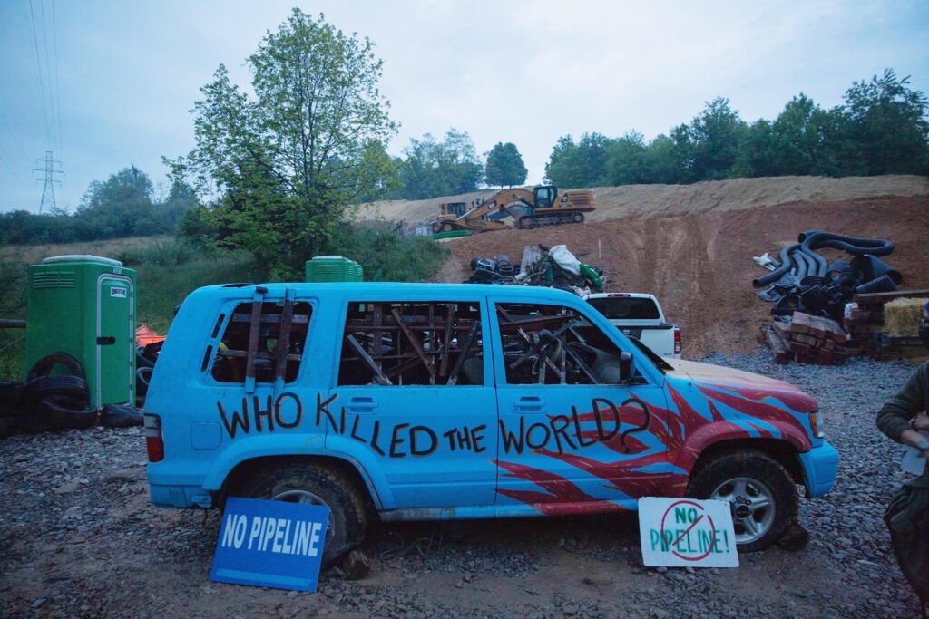 photo of an SUV deposited on an MVP development site, painted blue with flames from the front going backwards and the words "Who Killed the World?", with anti-MVP signs near the deflated tires